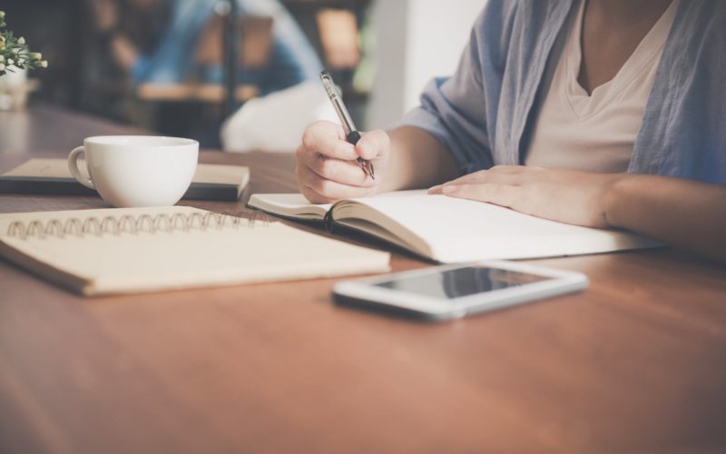 woman writing on a notebook beside teacup and tablet computer